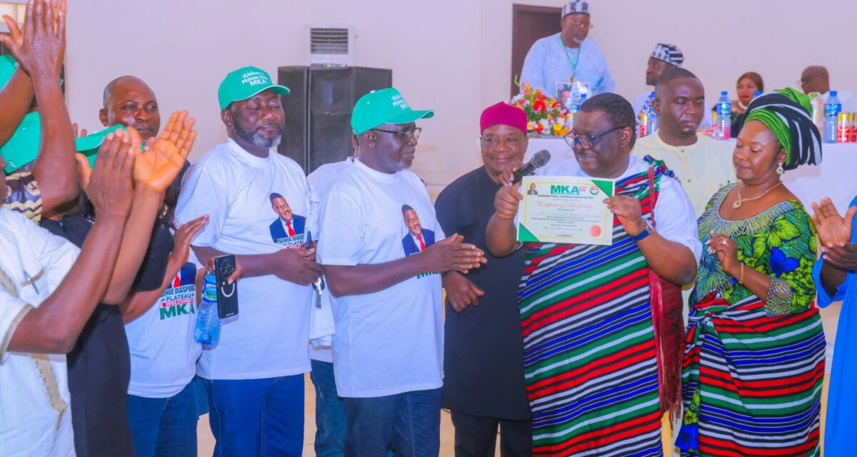 Man in a striped red, green, blue and white Tiv native wrap holds up a framed MKA 2027 certificate of membership while supporters in "Benue Diaspora Plateau Welcomes MKA" T-shirts and green caps applaud.