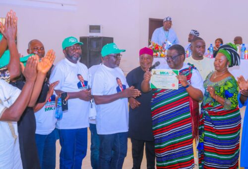 Man in a striped red, green, blue and white Tiv native wrap holds up a framed MKA 2027 certificate of membership while supporters in "Benue Diaspora Plateau Welcomes MKA" T-shirts and green caps applaud.