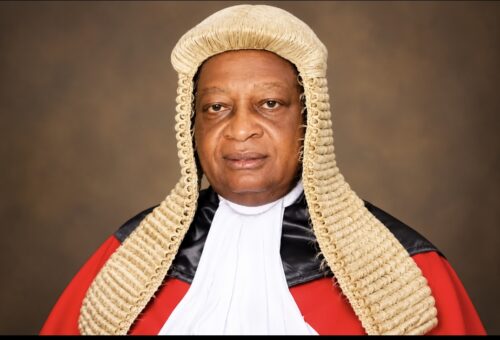Justice Maurice Ikpambese, Chief Judge of Benue State, in a judicial wig and red robe with white bands against a brown studio backdrop